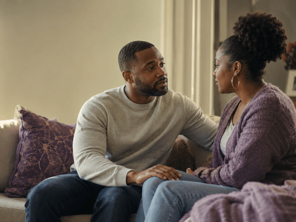 Black couple sitting on a couch facing each other in honest conversation about family patterns in their marriage